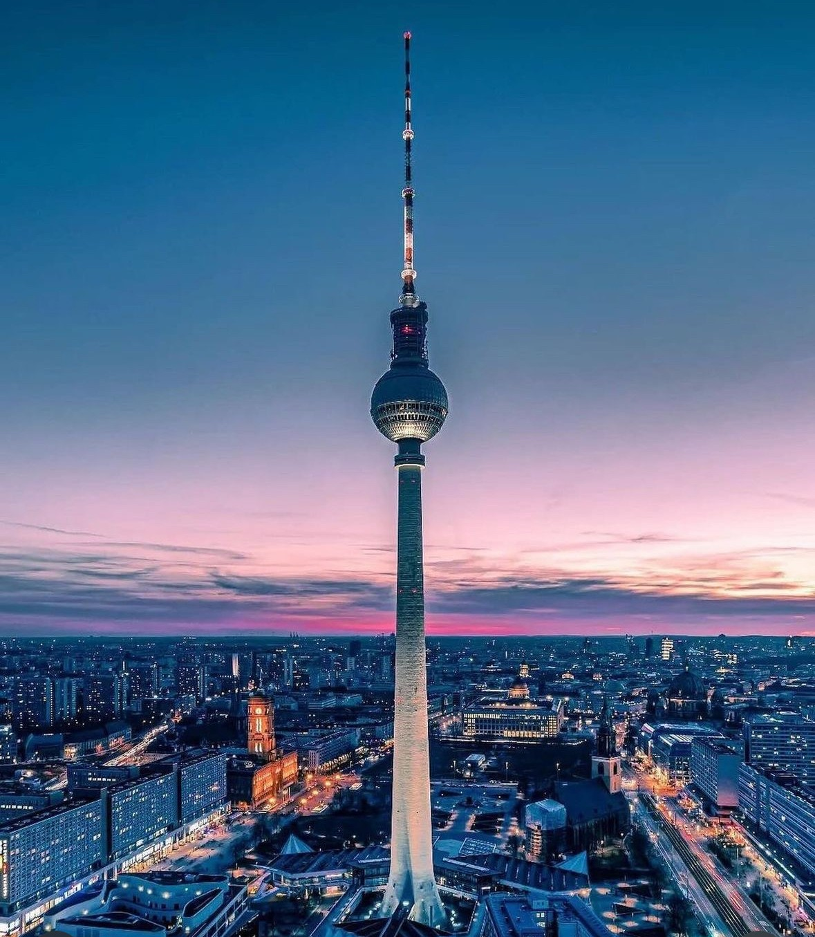 De Fernsehturm van Berlijn bij zonsondergang boven de skyline — de tv-toren steekt hoog uit boven de verlichte stad met een roze en blauwe hemel