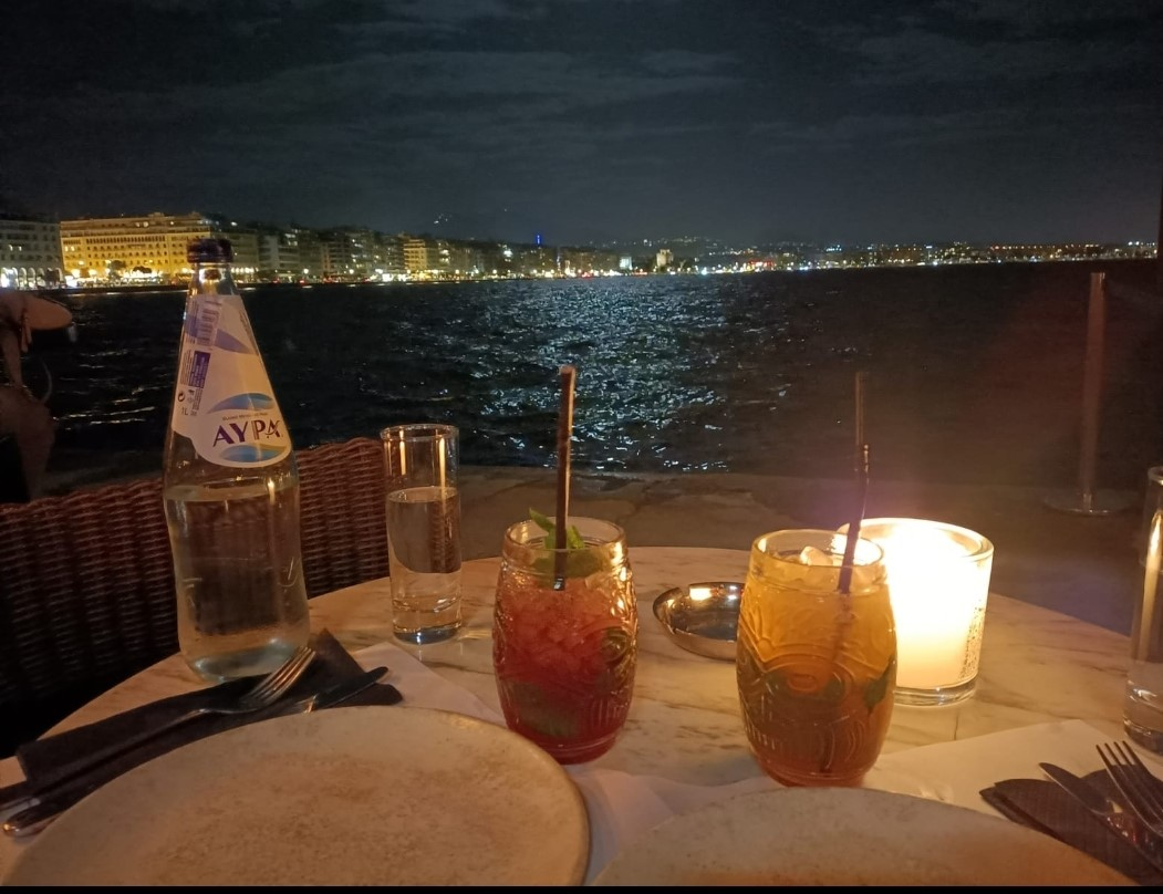 Nachtelijk uitzicht op de boulevard van Thessaloniki met stadslichten aan de overkant van het water, kaarslicht en glazen op een marmeren tafeltje