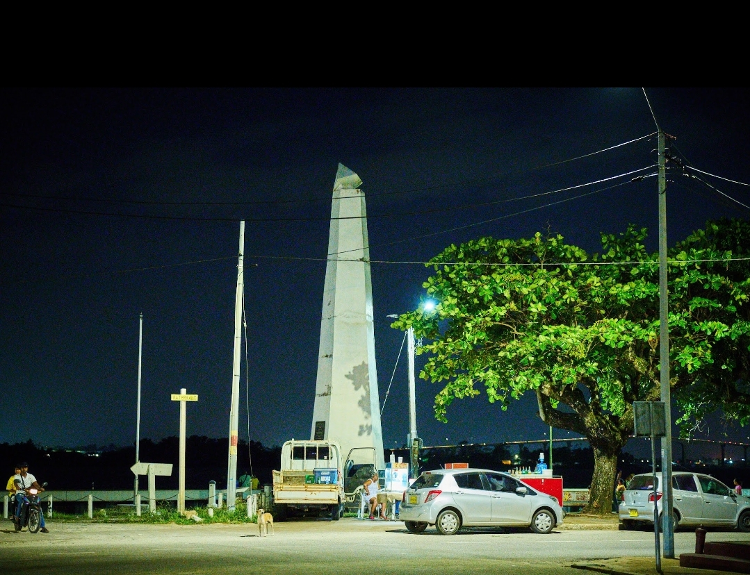 Waterkant Paramaribo bij nacht — obelisk verlicht in het donker met auto's, mensen en een vrachtwagen langs de kade