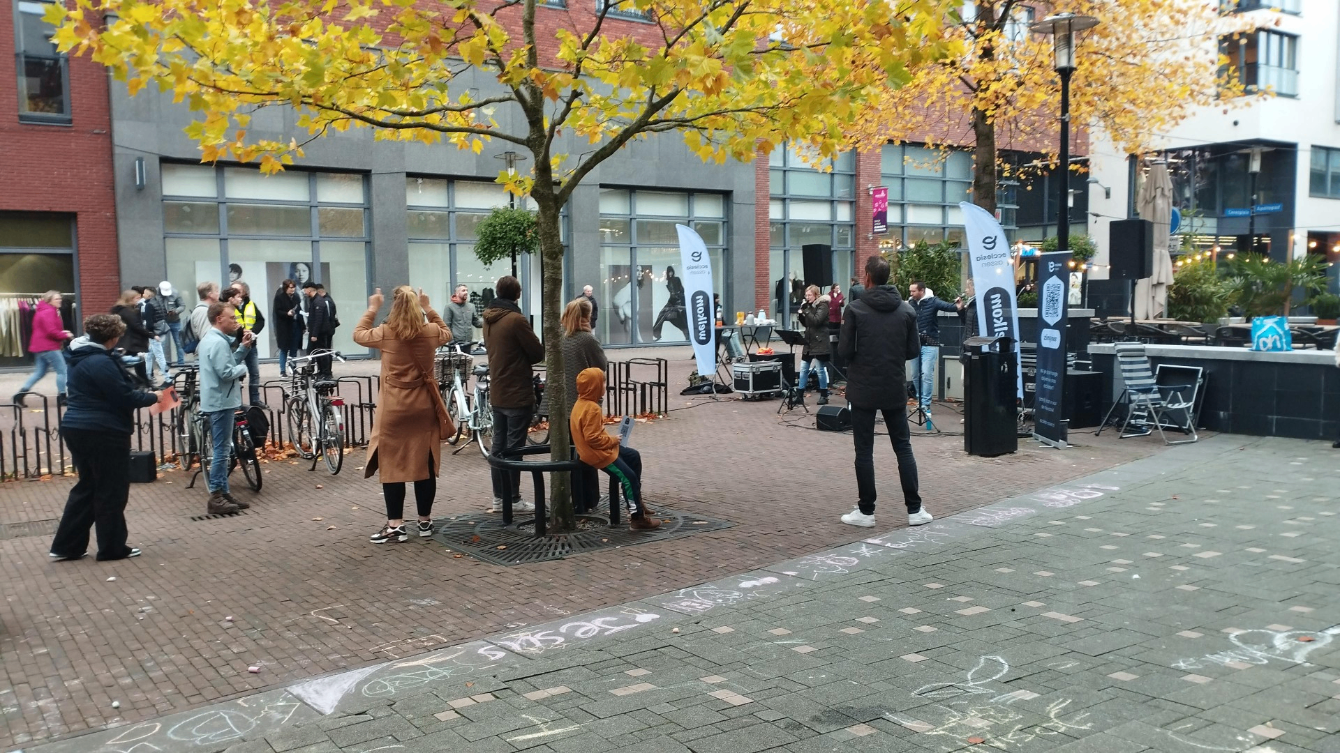 Evangelisatieactie op een plein in Drenthe — mensen luisteren naar aanbidding onder een herfstboom, krijttekeningen op de stoep