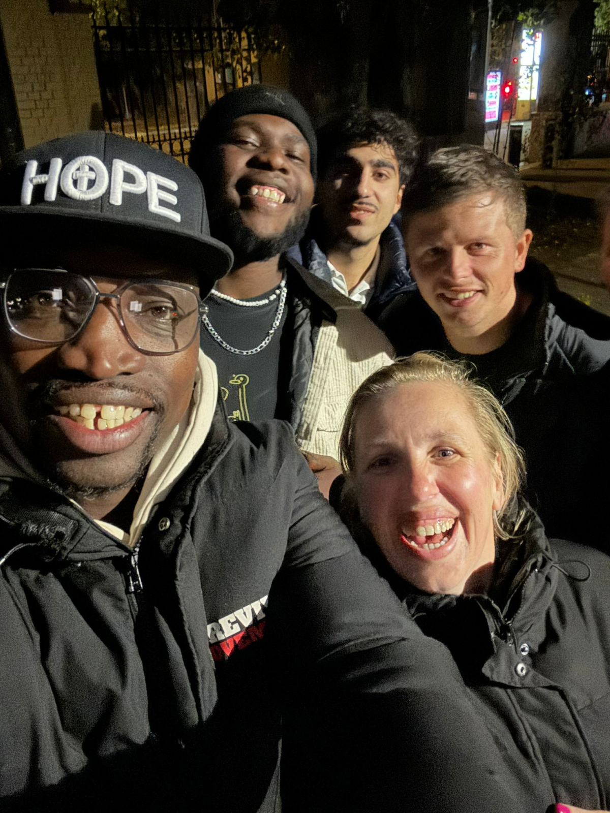 Selfie bij nacht in Berlijn — vijf mensen glimlachend samen, één met een pet met de tekst HOPE, na de ontmoeting met Eliyah en Misha