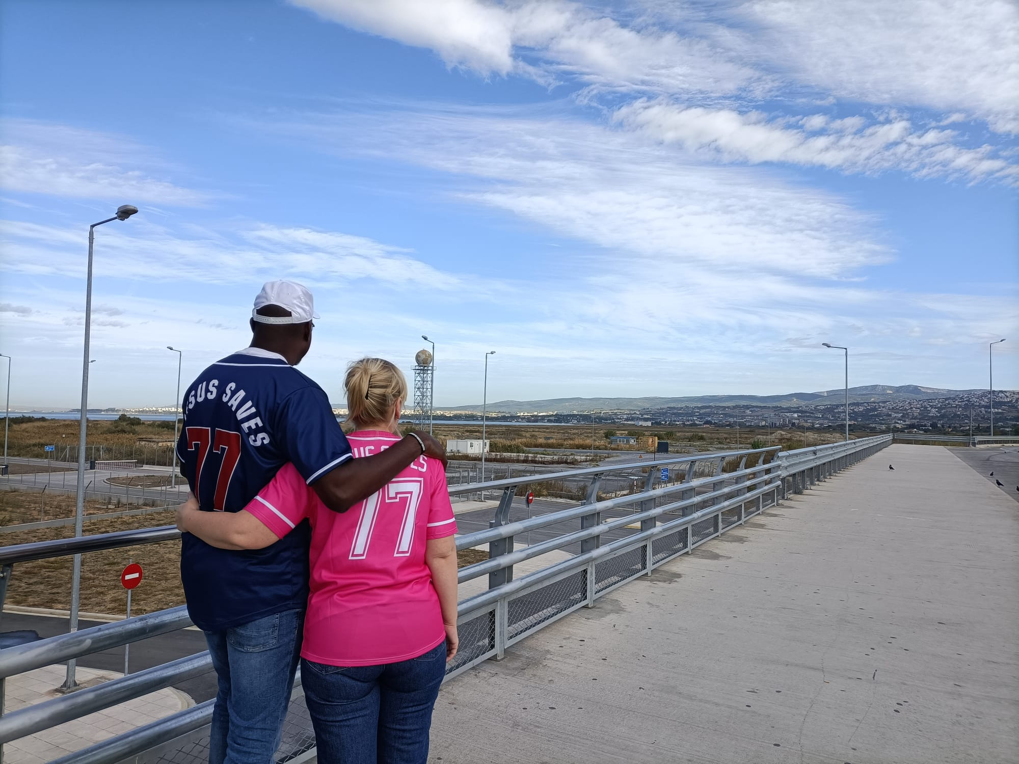 Clement en Lisa van Engel op de rug gezien, in Jesus Saves-shirts met het nummer 77, kijkend uit over een landschap met bergen en lucht